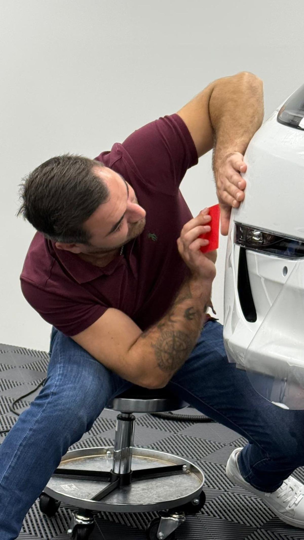 Mann in Jeans und Poloshirt auf Rollhocker bringt Schutzfolie an einem weißen Auto an.