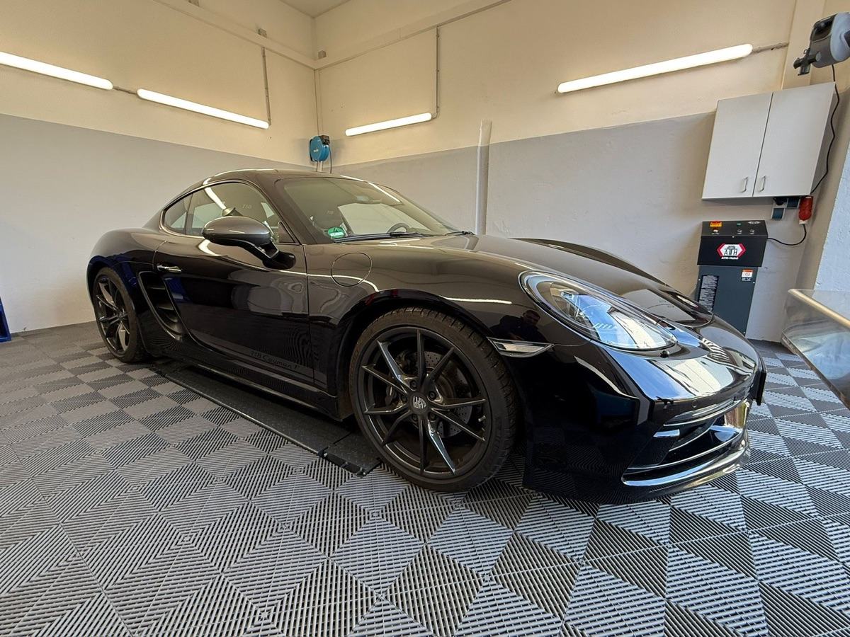 Schwarzer Porsche Cayman steht in einer Garage auf kariertem Boden.