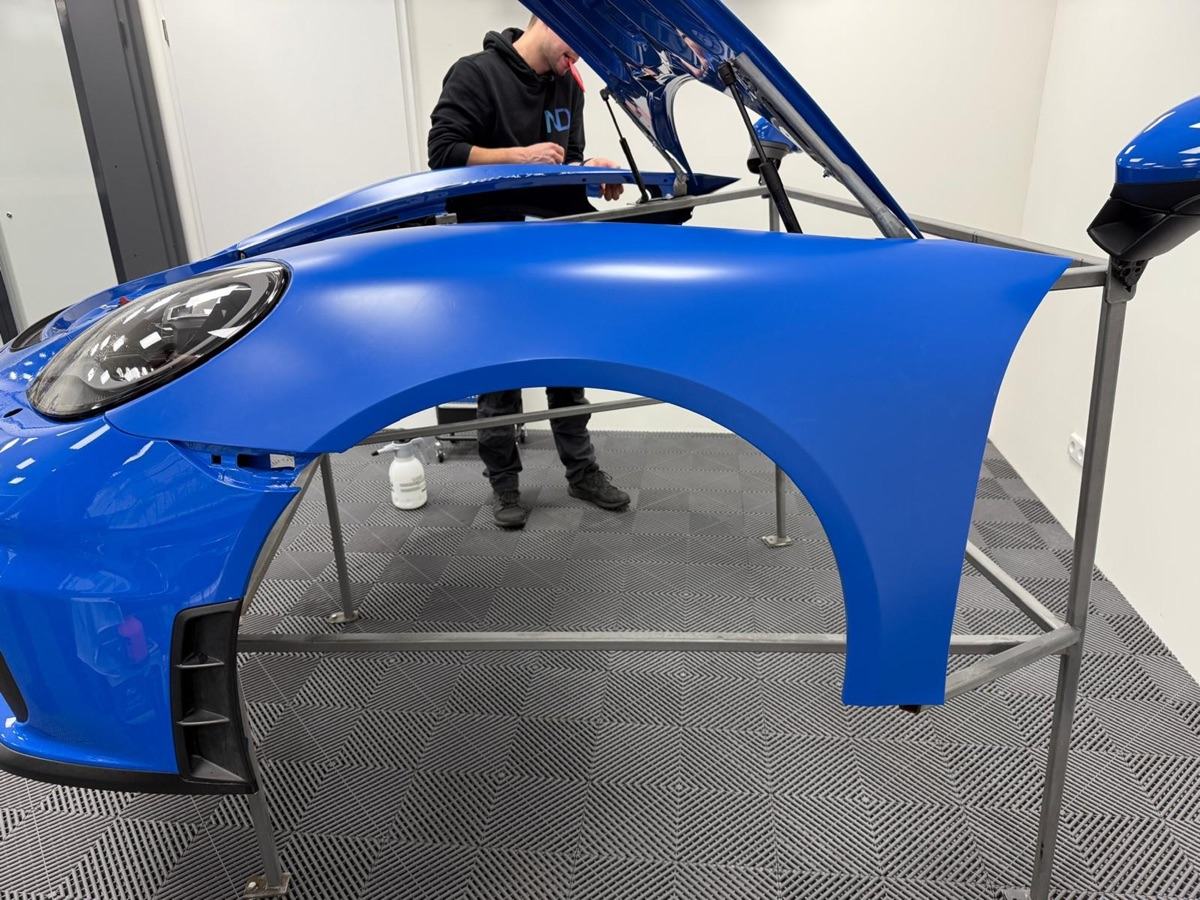 Blauer Porsche-Kotflügel und Motorhaube auf einem Metallgestell mit einer Person, die den Wagen foliert.