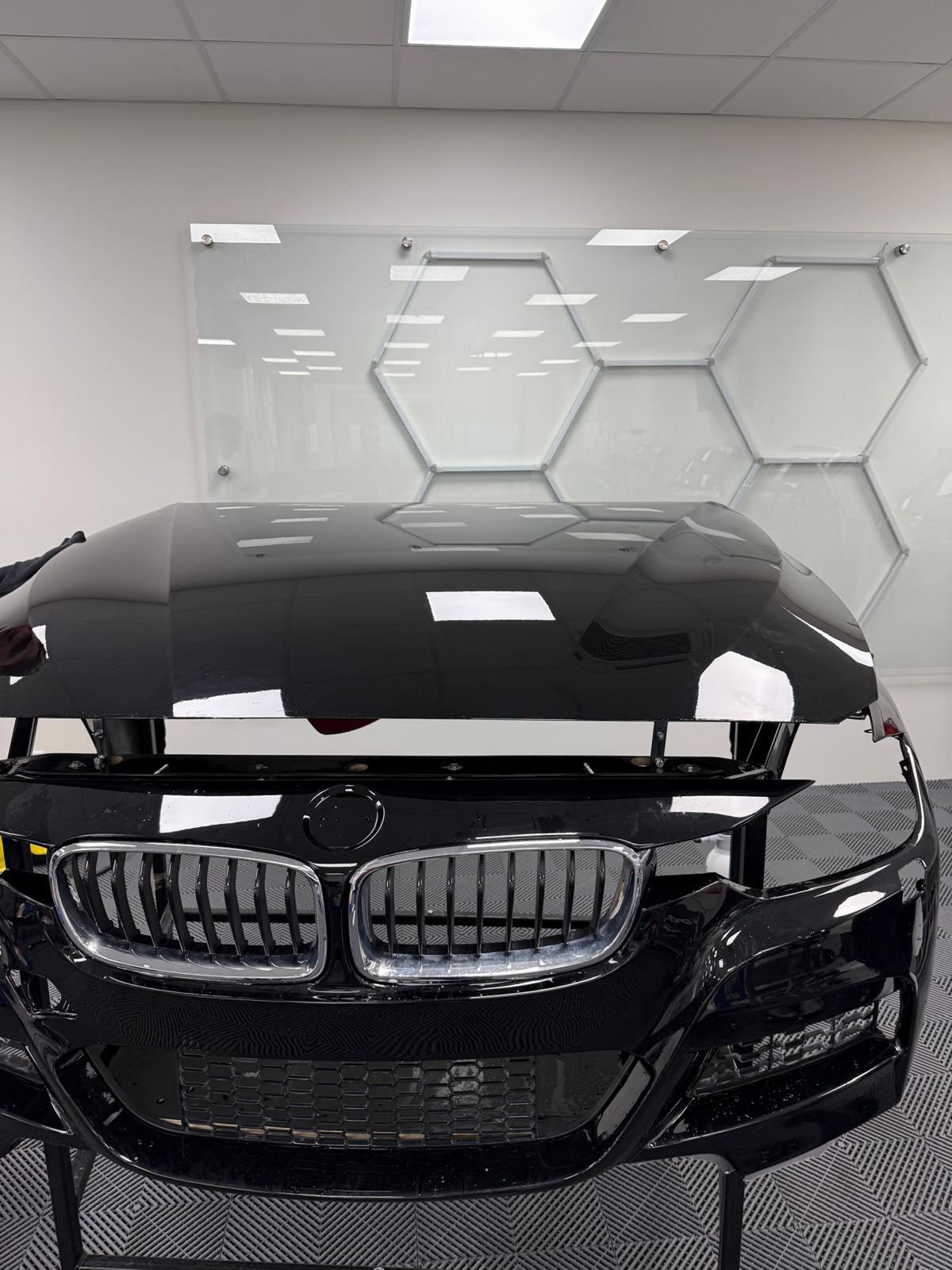 Schwarze BMW-Frontschürze mit glänzendem Kühlergrill und Motorhaube, reflektiert Deckenleuchten und sechseckige Wanddekoration.