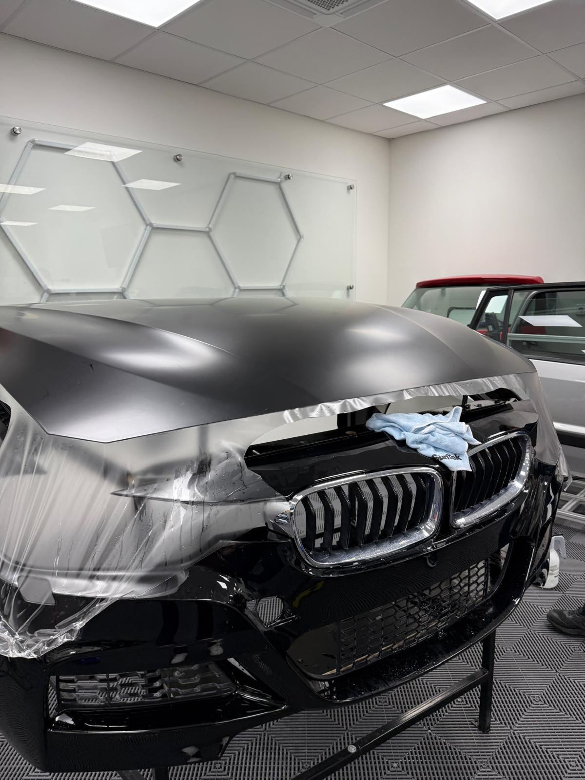 Schwarze BMW-Motorhaube und Stoßstange mit Schutzfolie und einem blauen Tuch in einem Raum mit sechseckigen Wandpaneelen.