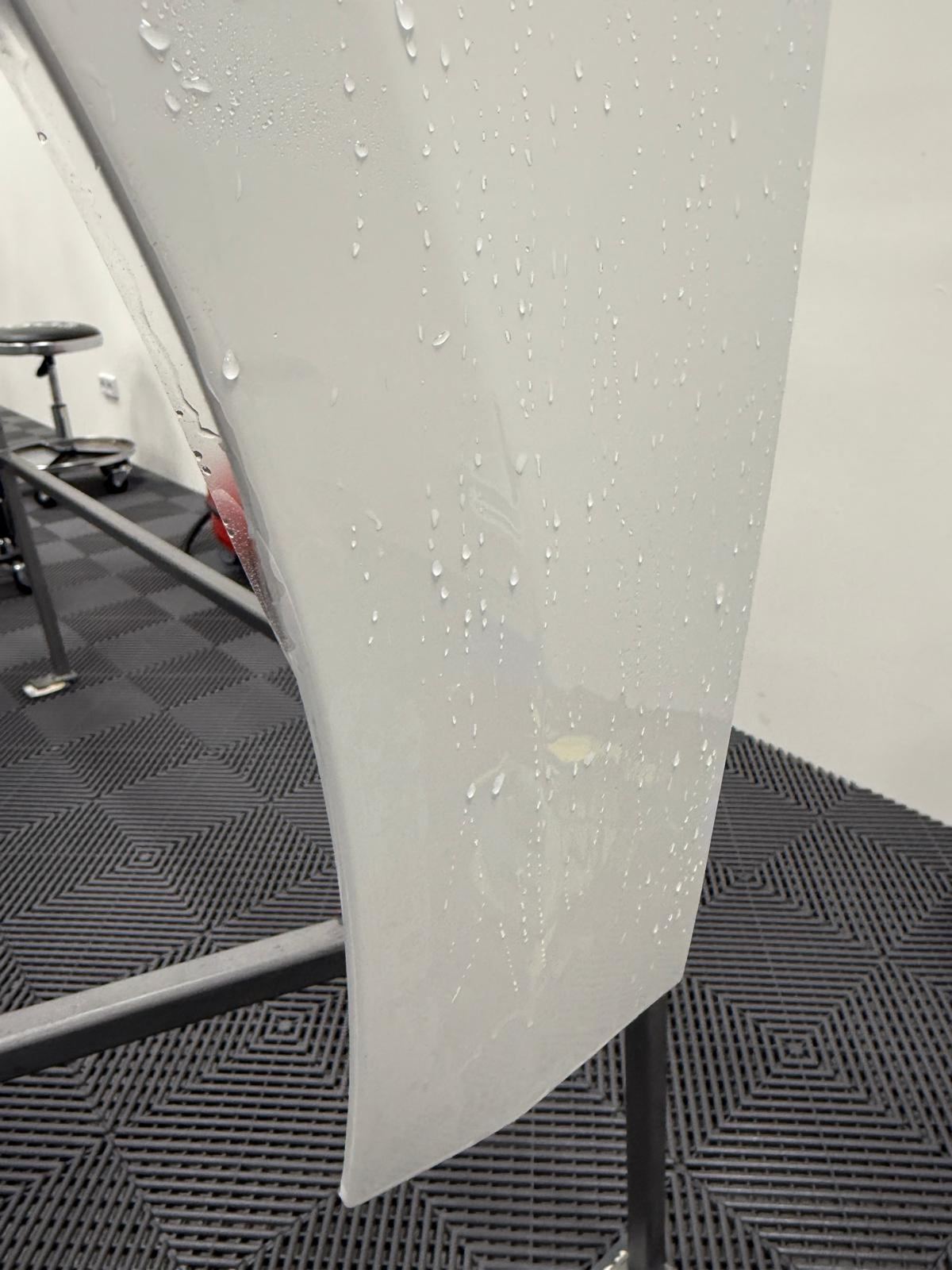 Weiße Autotür mit Wassertropfen auf einem grauen Gitterboden in einem Raum.
