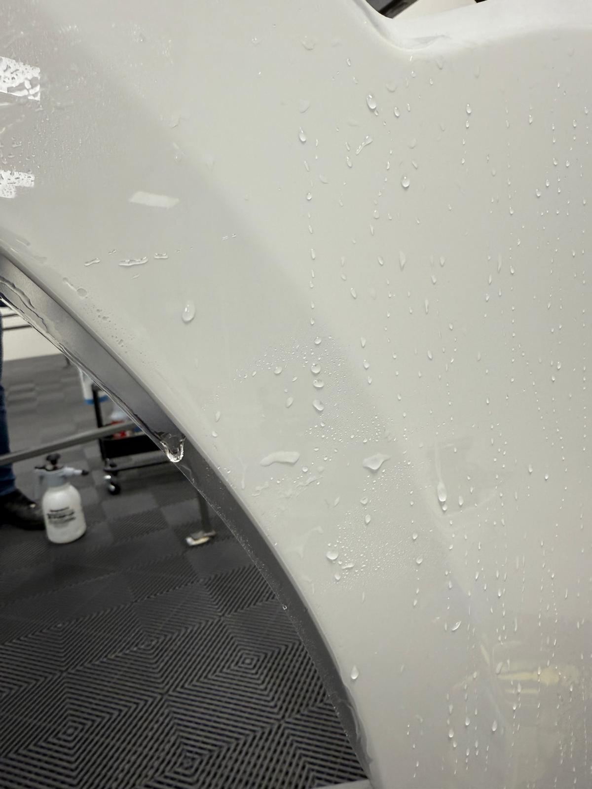 Weiße Autokarosserie mit Wassertropfen und Sprühflasche auf gemustertem Boden.